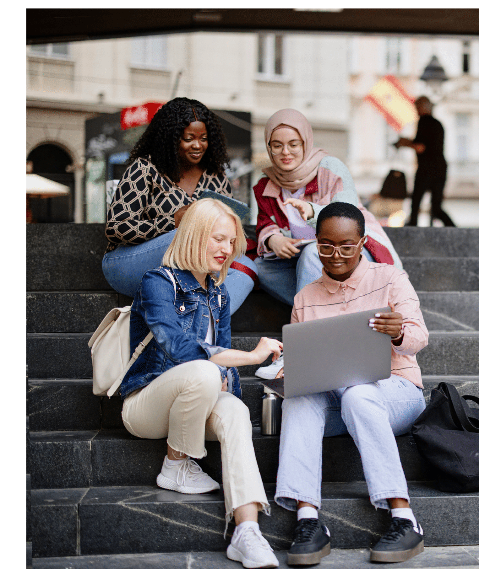 Four multiethnic girl friends walk around the city after or before the lecture. They wear casual clothes, holding electronic devices such as laptops, digital tablets or smartphones. They discuss lectures, talk and laugh, look at funny memes.