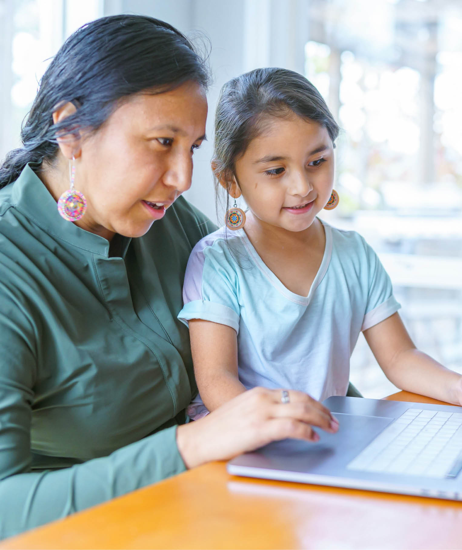 A Native American woman sits at the dining room table and works on a laptop computer as her adolescent son works on homework next to her. The woman's elementary age daughter is sitting on her lap and is looking at the computer screen.